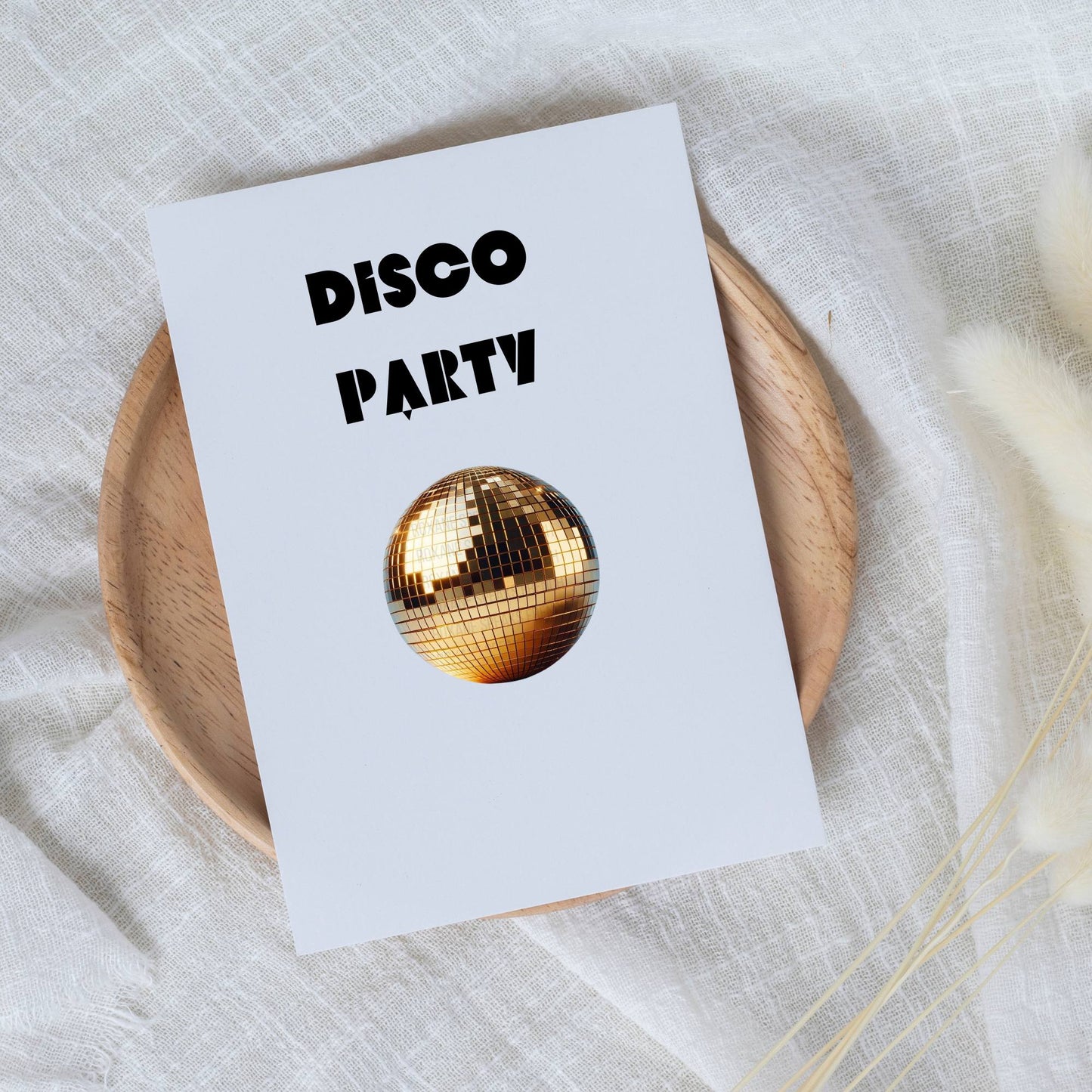 A party invitation card with "DISCO PARTY" printed in bold black letters, featuring a golden disco ball graphic at the center, all presented on a wooden plate against a delicate white fabric background.