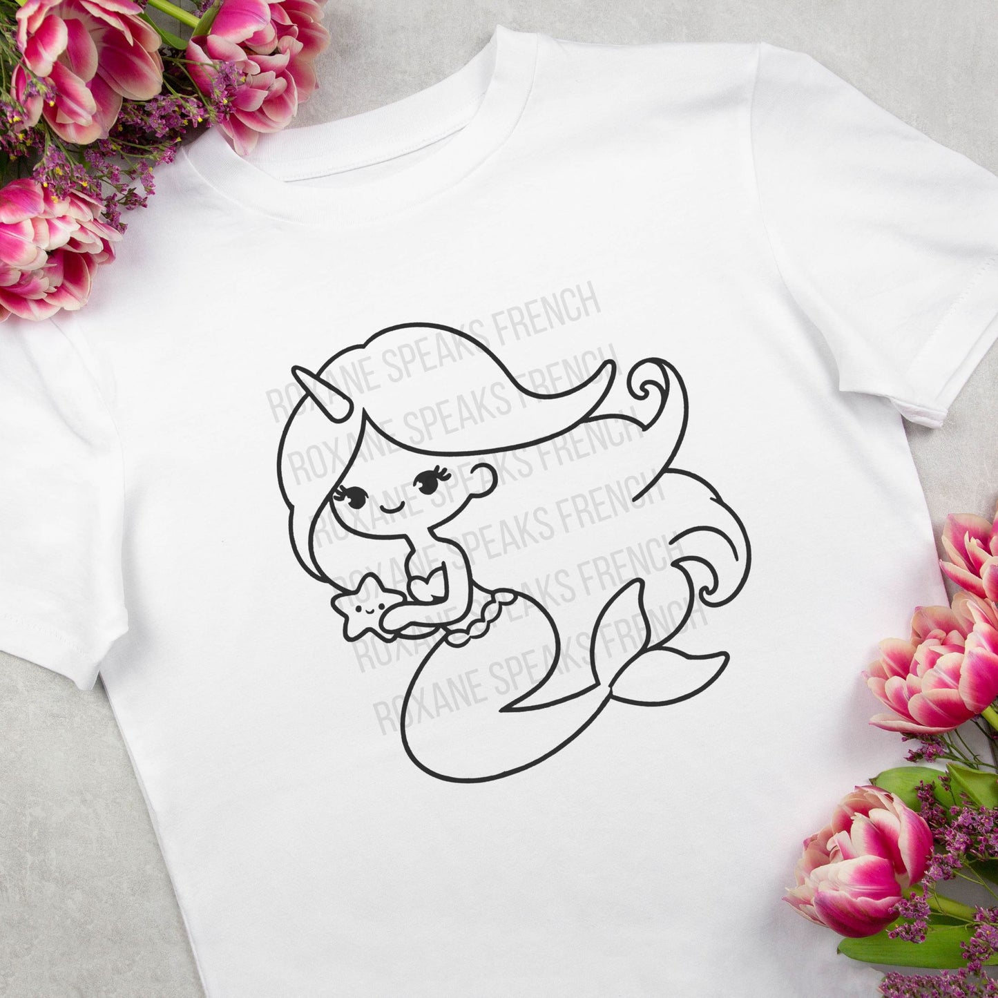 White t-shirt with a unicorn mermaid and starfish outline design, next to pink tulips.