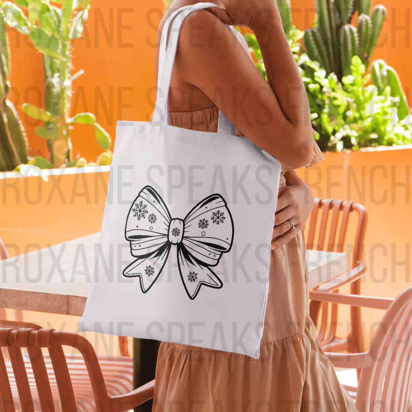 Person carrying a white tote bag with a Winter Bow SVG design, featuring an elegant bow adorned with snowflakes. The tote bag is showcased in a bright and cozy setting with plants and outdoor seating in the background.